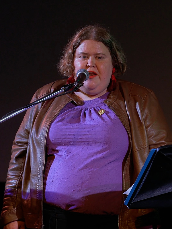 Alison Bergblom Johnson, a plus-sized woman with curly blonde hair wearing purple, large red earrings, and speaking into a microphone